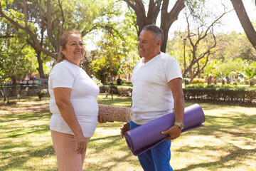 A senior man and woman stand facing each other, holding yoga mats under their arms, with contented...