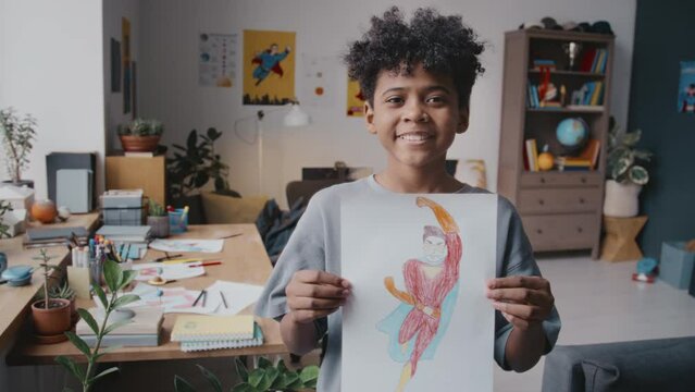 Portrait of little curly-haired African American boy holding superhero drawing and posing for camera with smile at home