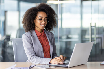 Side view of smiling hispanic woman with curly hair sitting at personal workplace and typing information on wireless laptop. Pretty lady using modern technologies for distance work in office interior.