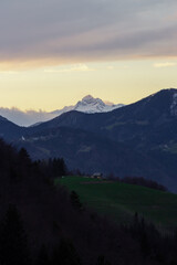 A three peaked mountain in the sunset with cloud cover over them and a small houde in front.