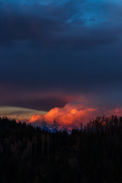 An amazing sunset over the mountains in orange colors with clouds hitting the peaks and rising up making a volcano effect. Forrest in the foreground of a golden hour lit mountain range in the back.
