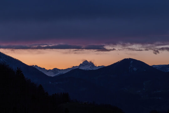 A three peaked mountain in the sunset with cloud cover over them and a small houde in front.