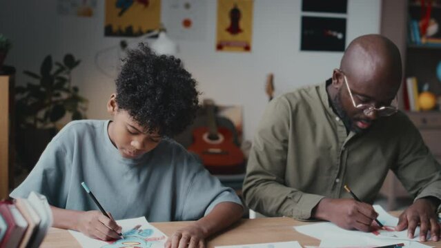 Black father giving advice to little son and praising him while drawing on paper with colored pencils together at home