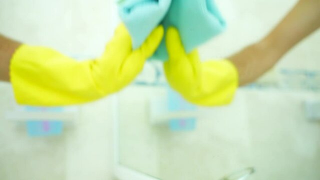 Close up of hand in a yellow rubber glove with blue rag cleaning the mirror in bathroom. Disinfect and sanitary standards