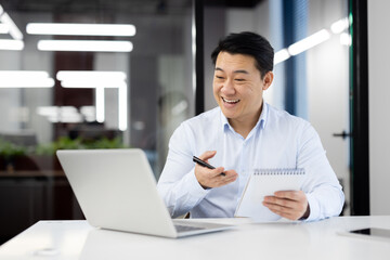 Smiling japanese male entepreneur having online video call on modern laptop while holding notebook and pen in hands. Positive man making notes during conversation with interlocutors at office.