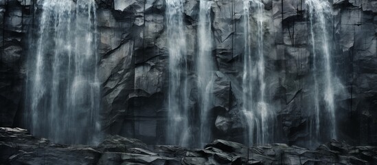 A monochrome photography of a waterfall cascading down a rocky cliff in a grey wooded natural landscape, with a bedrock and scattered rocks