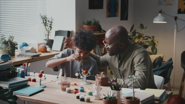 Cheerful African American man talking to little son and ruffling his hair while making DIY solar system model together at home