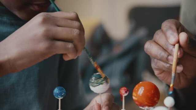 Close up tilt down shot of little African American kid sitting at desk and painting planet on DIY solar system model with assistance of father