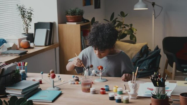 Little African American boy painting planets on DIY solar system model at desk in living room