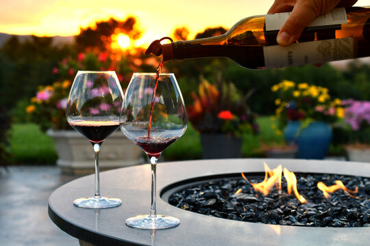 Pouring red wine into glasses at sunset golden hour next to a firepit fire table.