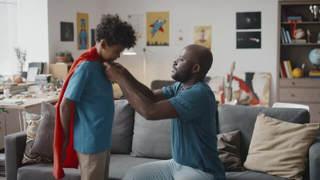 African American father putting red cape on little son, excited boy running around couch in living room while playing superhero at home