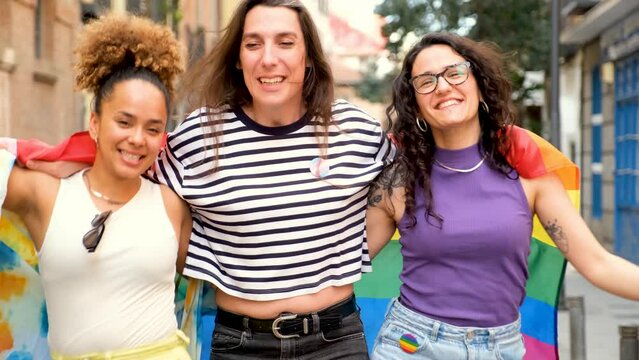 Three lesbian and trans women walking down the street wrapped in a rainbow flag. Scene is joyful and celebratory.