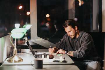 A dedicated man focuses on writing notes at his desk in an office during night time, exhibiting diligence and professionalism.