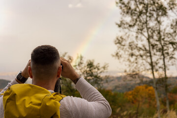 With a backpack on and wearing a sweater, a hiker gazes through binoculars, a rainbow visible in the distance following the rain.