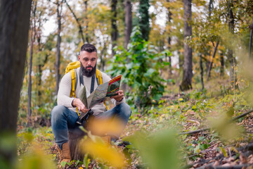 Naklejka na ściany i meble During his woodland hike, a hipster enjoys a rest, sitting on a tree stump while consulting a paper map to plan his route.