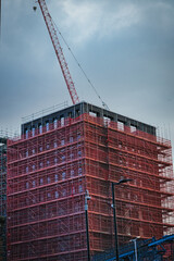 Construction site with a tall building covered in red scaffolding and a crane against a cloudy sky...