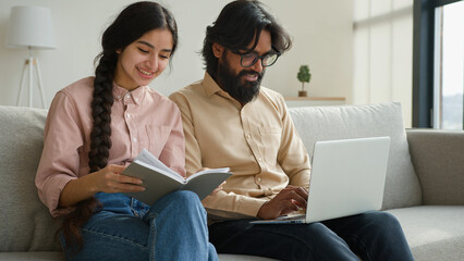 Diverse couple on couch with laptop pay bills loan payments planning household budget arabian woman wife read notes in notebook indian man husband typing data email on computer family business at home