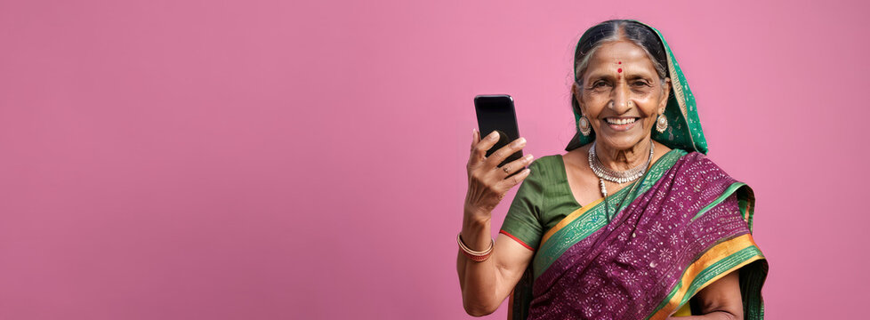 Elderly smiling Indian woman with grey hair and bindi holding mobile phone isolated on pink studio background, wide banner for advertising with copy space for text