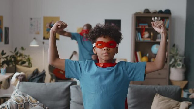 Portrait of little African American boy in superhero costume showing biceps and posing for camera while his dad running in background at home