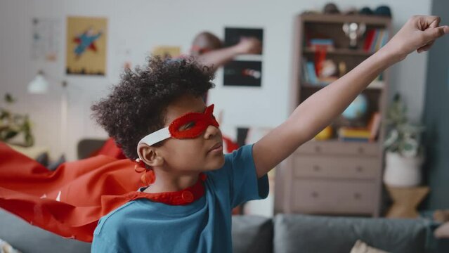 Portrait of little Black boy in mask standing with one arm raised and red cape fluttering in wind, turning to camera and smiling while imitating superhero flight at home, dad running behind
