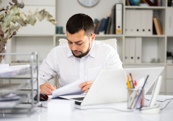 European man office worker sitting at table in front of his laptop and making notes on paper.