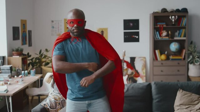 Slow motion shot of adult African American man in superhero mask and red cape spinning around and posing for camera with arms crossed in living room at home