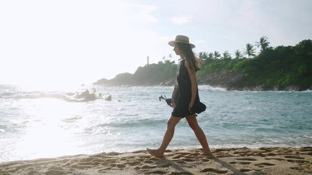 Creative woman in dress, hat carries tripod, strolls along sandy beach. Artist walks by sea, waves crash on shore. Content creator films at coastal location. Sunlit tropical scenery, travel blogger.