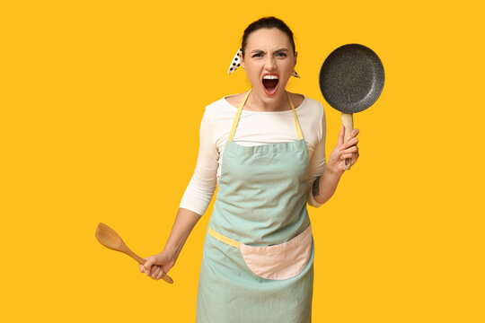 Portrait of angry young housewife in apron with frying pan and wooden spatula on yellow background