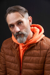 A man with a beard and gray hair is wearing a brown jacket and orange hoodie. He is looking at the camera with a serious expression