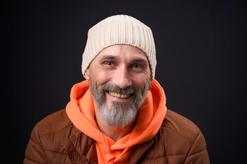 A man wearing a brown jacket and orange hoodie is smiling. He has a white knit hat on his head
