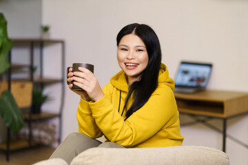 A woman in a yellow hoodie is smiling while holding a mug. The room has a cozy atmosphere with a couch and a potted plant