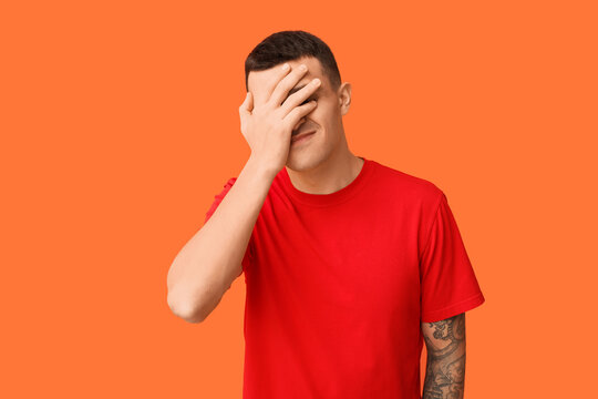 Handsome ashamed young man covering face with hand on orange background