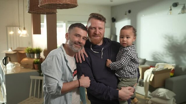 Portrait Of A Happy Gay Couple With Their Young Son At Home With Moving Boxes In The Background