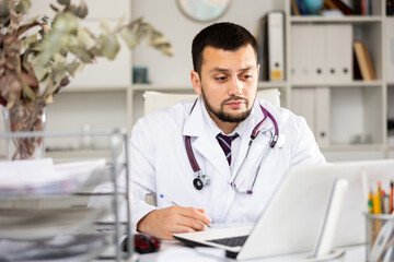 Portrait of skillful man physician sitting at table in his office and writing.