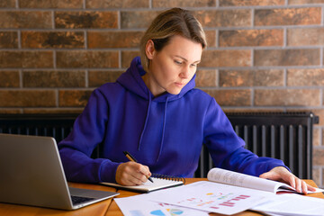 A woman in a blue sweater is working at a desk, writing notes in a notebook, reading a book. Female student studying, doing assignments, laptop on table, open textbook, space for copy