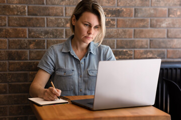 A young girl works on a laptop and writes notes in a notebook. Business Woman Blonde In Blue Shirt In Office, Girl In Cafeteria Studying Making Report, Writing Book, Making Business Plan
