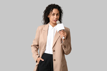 Beautiful young African-American female journalist with microphone on grey background