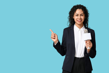 Beautiful young happy African-American female journalist with microphone pointing at something on blue background