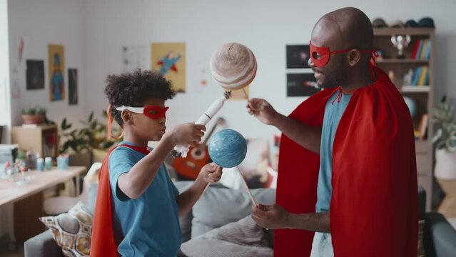 Little African American kid and his dad wearing superhero costumes standing in living room and playing with toy planets and rocket while having fun together at home