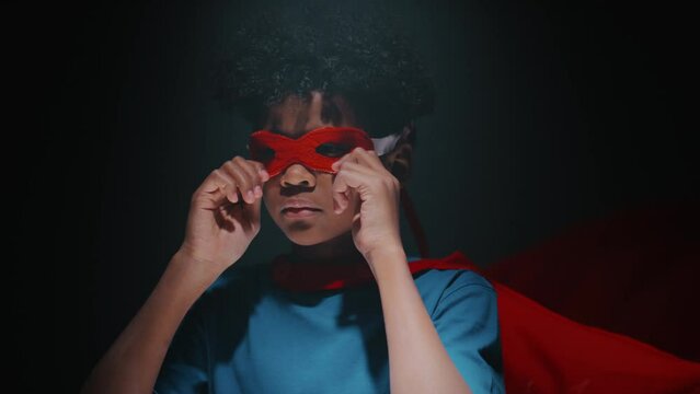 Portrait of little African American boy in red cape putting superhero mask on face, folding his arms and posing for camera with confidence against dark background in studio