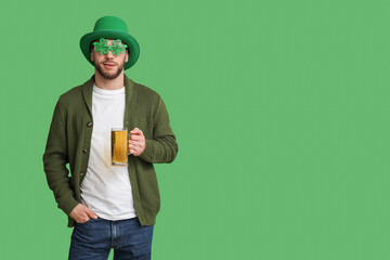 Young man in leprechaun hat and decorative glasses in shape of clover with glass of beer on green background. St. Patrick's Day celebration