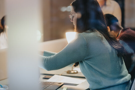 A concentrated female professional is intensely working on her laptop at an office desk, with coworkers around her.