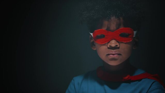 Portrait of little African American boy in superhero costume turning to camera and posing with confidence against black background in studio