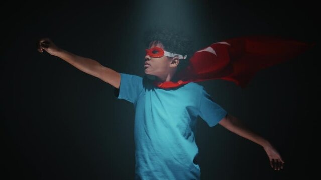 Little Black boy in superhero mask and red cape fluttering in wind standing with one arm straight out and imitating flight while posing in studio against dark background