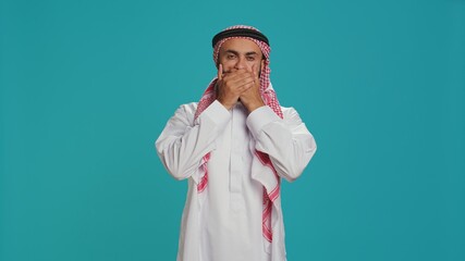 Muslim adult covers eyes, ears and mouth in studio, showcasing three wise monkeys metaphor sign. Middle eastern guy presenting dont hear, see or speak concept symbol in arabic clothes.