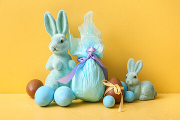 Painted and chocolate Easter eggs wrapped in paper with toy bunnies on yellow background
