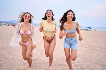 Three cheerful young swimwear women enjoying running excited along beach. Caucasian fitness girls celebrating summer vacation together outdoors. Female friends people having fun sunny weekend holiday