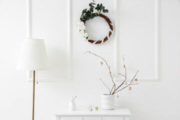 Vase with branches on table, floor lamp and Easter wreath near white wall