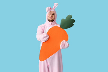 Young man in bunny costume with paper carrot on blue background. Easter celebration