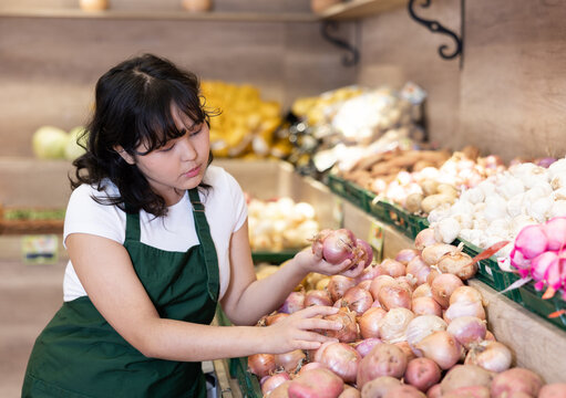 Attentive young saleswoman placing fresh onions on food stall in grocery store
